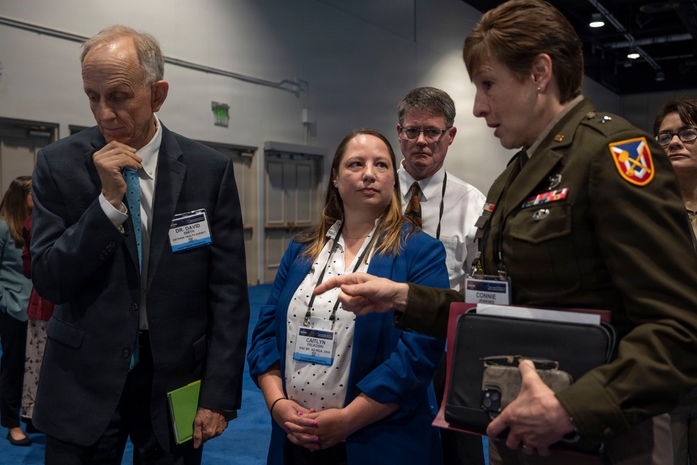 A group of professionals are gathered at what appears to be a conference or professional event. The group includes a military officer in Army dress uniform, civilian attendees with name badges, and appears to involve a briefing or discussion. Name badges visible include 'Dr. David' affiliated with Defense Health Agency and 'Caitlyn Felkosan' affiliated with PAE.