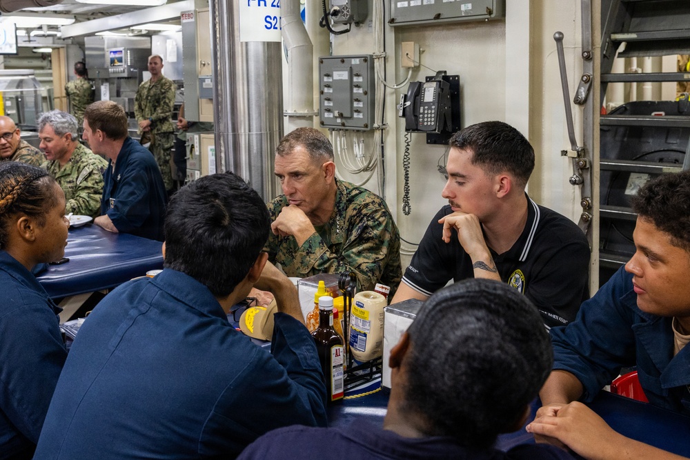 Military personnel are gathered in what appears to be a ship's mess hall or dining facility, engaged in conversation around tables. A Marine in camouflage uniform sits among sailors wearing navy blue coveralls, suggesting a cross-branch interaction or briefing. The setting includes institutional fixtures such as a wall-mounted telephone, electrical panels, and conduit piping typical of a naval vessel.