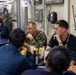 Military personnel are gathered in what appears to be a ship's mess hall or dining facility, engaged in conversation around tables. A Marine in camouflage uniform sits among sailors wearing navy blue coveralls, suggesting a cross-branch interaction or briefing. The setting includes institutional fixtures such as a wall-mounted telephone, electrical panels, and conduit piping typical of a naval vessel.