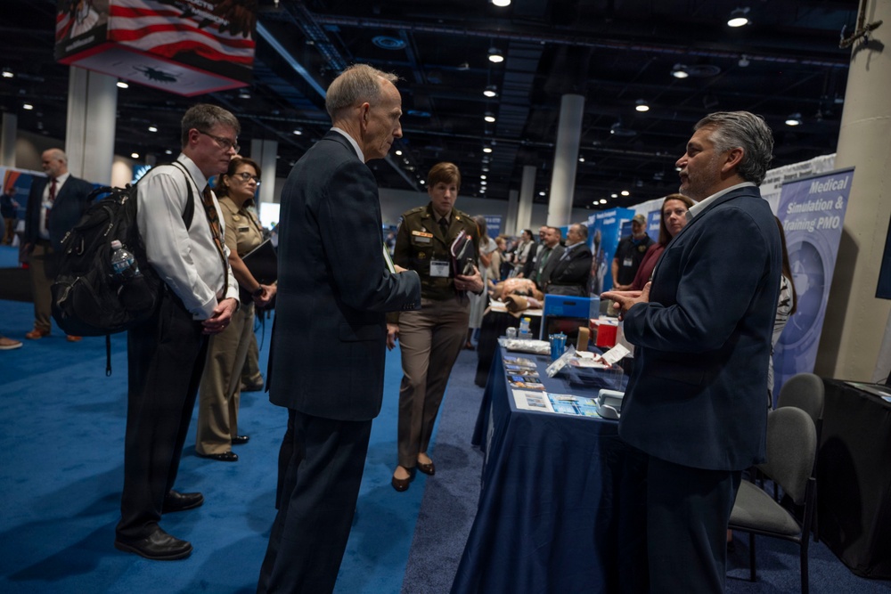 A professional conference or trade show floor featuring military and civilian personnel interacting at exhibition booths. The visible signage references 'Medical Simulation &amp; Training PMO,' suggesting a defense or military medical technology event. Attendees in both military uniforms and civilian business attire are engaged in conversations at display tables.