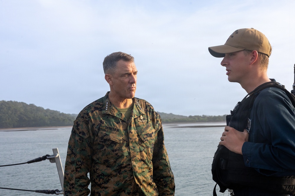 Two individuals are engaged in conversation aboard what appears to be a military or patrol vessel on a body of water. The person on the left is wearing U.S. Marine Corps MARPAT camouflage uniform with a name tape reading 'DONOVAN' and general officer stars visible. The person on the right is wearing civilian-style clothing with a tan baseball cap and a tactical vest.