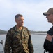 Two individuals are engaged in conversation aboard what appears to be a military or patrol vessel on a body of water. The person on the left is wearing U.S. Marine Corps MARPAT camouflage uniform with a name tape reading 'DONOVAN' and general officer stars visible. The person on the right is wearing civilian-style clothing with a tan baseball cap and a tactical vest.