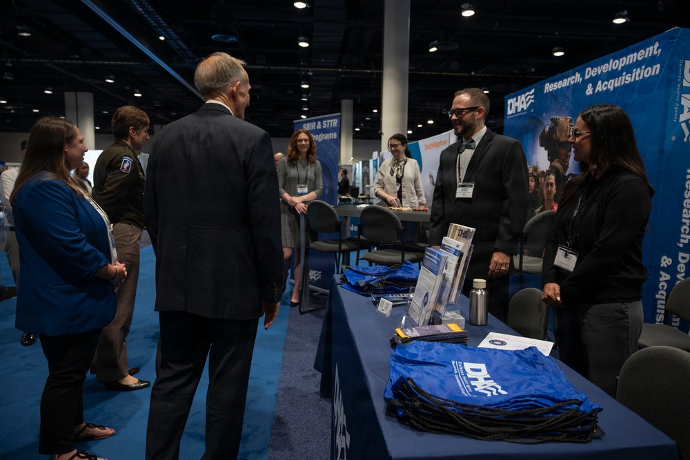 A professional conference or trade show event featuring a DHA (Defense Health Agency) exhibit booth focused on Research, Development, and Acquisition. Several attendees and booth representatives are engaged in conversation around a display table covered with branded materials, brochures, and blue DHA drawstring bags. The setting appears to be a large convention center with multiple exhibitor booths visible in the background.