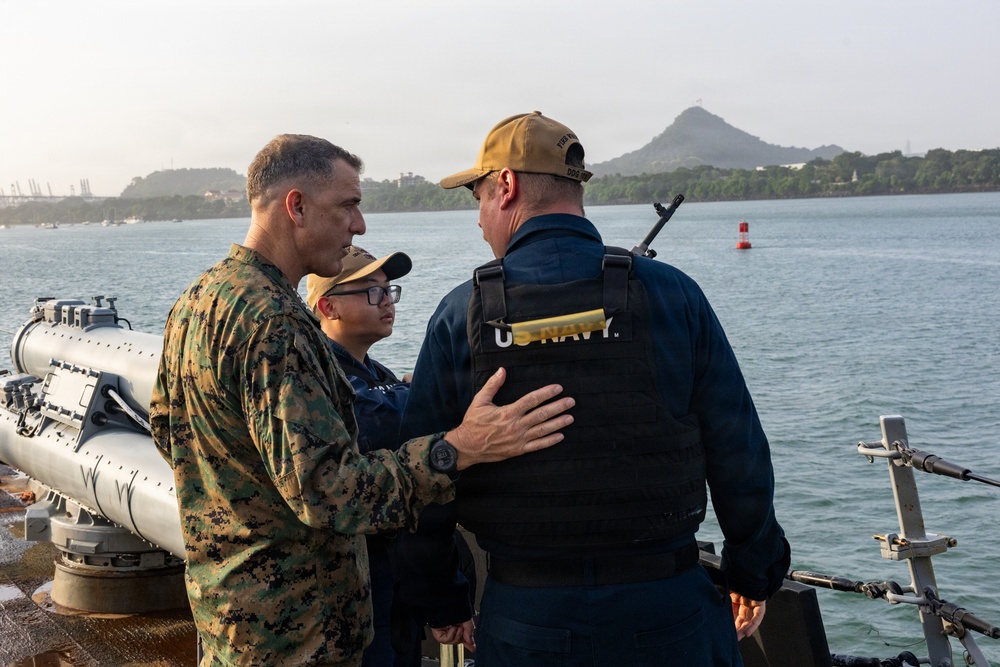Three military personnel are standing on the deck of a naval vessel, engaged in conversation. One individual is wearing U.S. Marine Corps camouflage utilities, one is in U.S. Navy working uniform with a tactical vest marked 'U.S. NAVY', and a third smaller individual stands between them. The scene appears to take place in a harbor or waterway with a distinctive hill or small mountain visible in the background.