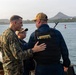 Three military personnel are standing on the deck of a naval vessel, engaged in conversation. One individual is wearing U.S. Marine Corps camouflage utilities, one is in U.S. Navy working uniform with a tactical vest marked 'U.S. NAVY', and a third smaller individual stands between them. The scene appears to take place in a harbor or waterway with a distinctive hill or small mountain visible in the background.