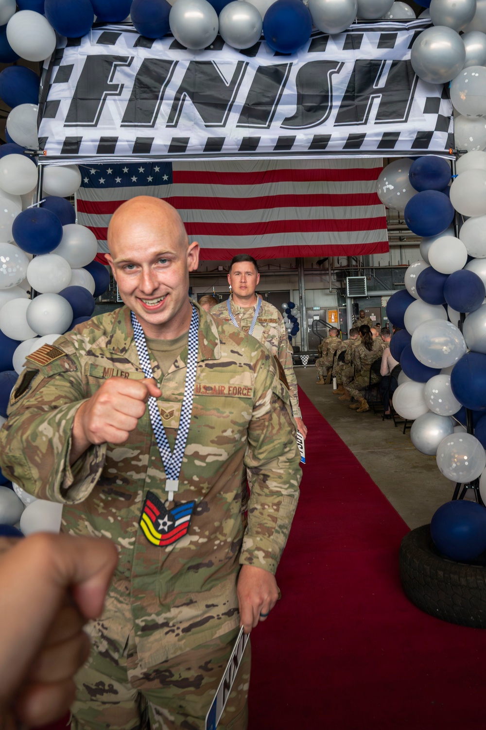A U.S. Air Force service member named Miller, wearing OCP (Operational Camouflage Pattern) uniform with a Staff Sergeant rank patch, crosses under a 'FINISH' arch decorated with blue, silver, and white balloons while wearing a medal with a checkered lanyard. An American flag is visible in the background along with other uniformed personnel also wearing medals. The setting appears to be an indoor military hangar hosting a ceremonial or competitive event.