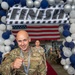 A U.S. Air Force service member named Miller, wearing OCP (Operational Camouflage Pattern) uniform with a Staff Sergeant rank patch, crosses under a 'FINISH' arch decorated with blue, silver, and white balloons while wearing a medal with a checkered lanyard. An American flag is visible in the background along with other uniformed personnel also wearing medals. The setting appears to be an indoor military hangar hosting a ceremonial or competitive event.