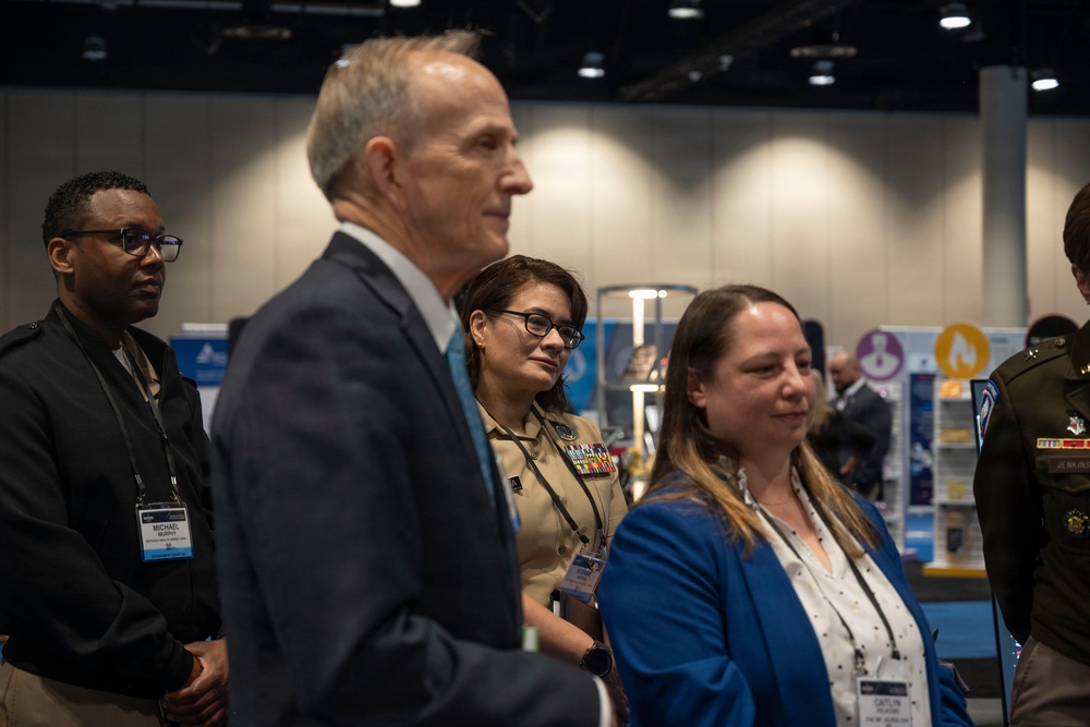 A group of professionals attending what appears to be a conference or expo, standing together in an exhibition hall. The group includes civilians and at least two individuals in military uniforms, all wearing name badge lanyards. They appear to be listening to a presentation or observing exhibits in the background.