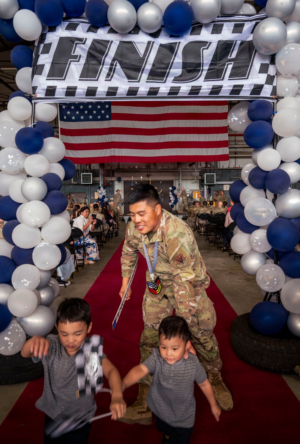 A military service member in camouflage uniform walks through a celebratory 'FINISH' arch decorated with blue, white, and silver balloons, accompanied by two young children on a red carpet. An American flag is displayed prominently in the background, and the event appears to be a military graduation or deployment return ceremony held inside a hangar. The soldier is smiling and holding what appears to be a diploma or certificate while guiding the children forward.