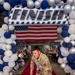 A military service member in camouflage uniform walks through a celebratory 'FINISH' arch decorated with blue, white, and silver balloons, accompanied by two young children on a red carpet. An American flag is displayed prominently in the background, and the event appears to be a military graduation or deployment return ceremony held inside a hangar. The soldier is smiling and holding what appears to be a diploma or certificate while guiding the children forward.