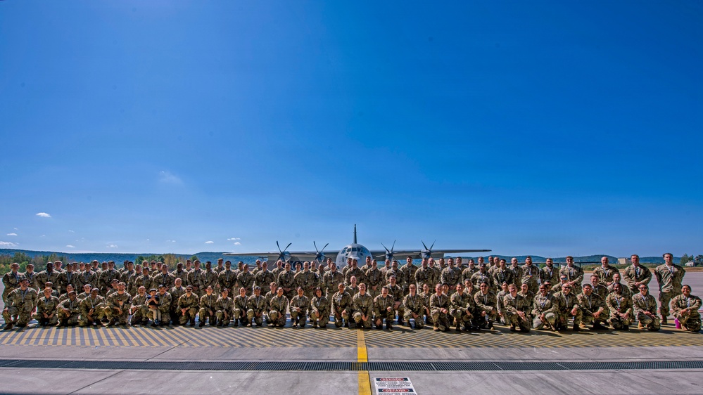 A large group of military personnel in camouflage uniforms are posed for a formal group photograph on an airfield tarmac. Behind them, centered in the frame, is a four-engine turboprop military transport aircraft, consistent with a C-130 Hercules. The photo appears to be taken on a clear, sunny day at a military air base.