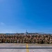 A large group of military personnel in camouflage uniforms are posed for a formal group photograph on an airfield tarmac. Behind them, centered in the frame, is a four-engine turboprop military transport aircraft, consistent with a C-130 Hercules. The photo appears to be taken on a clear, sunny day at a military air base.