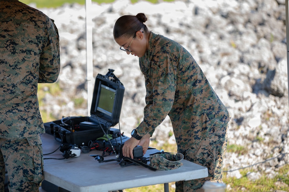 Two military personnel wearing MARPAT (Marine pattern) camouflage uniforms are working at a folding table set up outdoors under a canopy. The central figure, wearing glasses and hair in a bun, is leaning over the table handling electronic equipment. The setting appears to be a field operations site with rocky terrain visible in the background.
