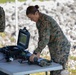 Two military personnel wearing MARPAT (Marine pattern) camouflage uniforms are working at a folding table set up outdoors under a canopy. The central figure, wearing glasses and hair in a bun, is leaning over the table handling electronic equipment. The setting appears to be a field operations site with rocky terrain visible in the background.