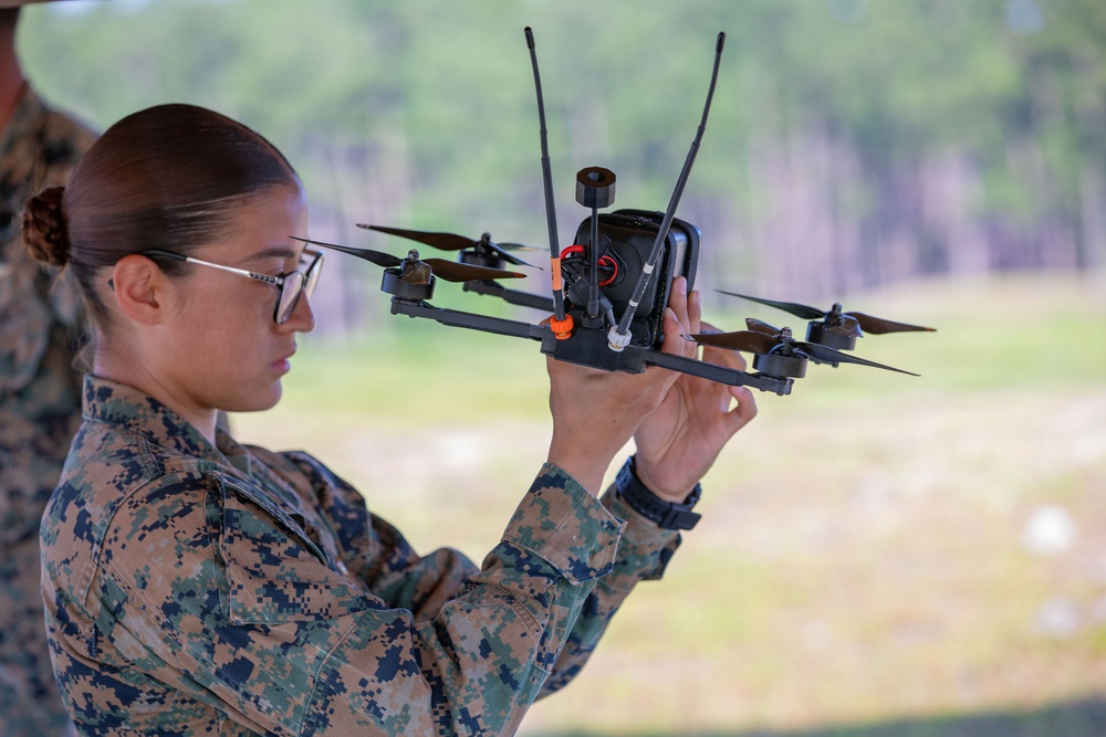 A female military service member in MARPAT (Marine pattern) camouflage uniform is closely inspecting or preparing a quadcopter FPV (First Person View) drone. She is holding the drone at eye level, examining it carefully in what appears to be an outdoor training environment. The setting suggests a military drone training or operational exercise.