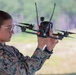A female military service member in MARPAT (Marine pattern) camouflage uniform is closely inspecting or preparing a quadcopter FPV (First Person View) drone. She is holding the drone at eye level, examining it carefully in what appears to be an outdoor training environment. The setting suggests a military drone training or operational exercise.