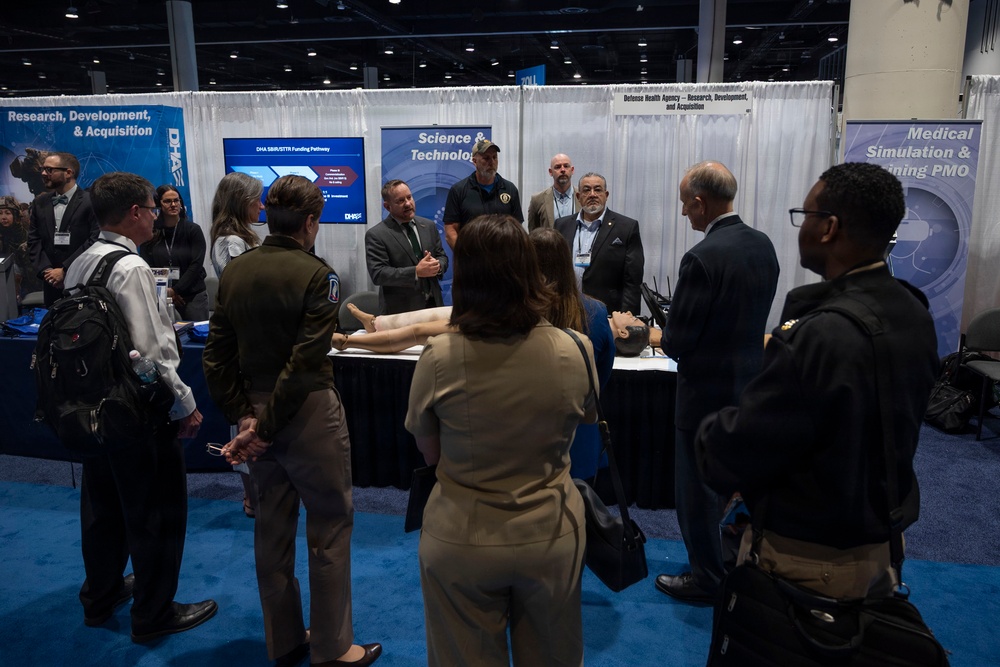 A group of military and civilian personnel are gathered at a Defense Health Agency (DHA) exhibition booth at what appears to be a professional conference or trade show. The booth displays banners for 'Research, Development, &amp; Acquisition', 'Science &amp; Technology', and 'Medical Simulation &amp; Training PMO'. Attendees are viewing medical simulation mannequins and a presentation screen showing 'DHA SBIR/STTR Funding Pathway'.