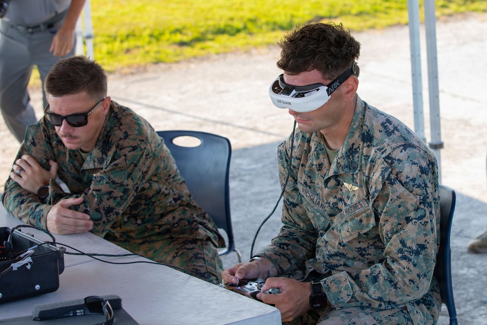 Two U.S. Marines in MARPAT digital camouflage uniforms are seated at a table under a canopy, operating what appears to be a drone or unmanned aerial system. The Marine on the right is wearing FPV (First Person View) goggles branded 'ARK' and holding a remote controller, while the Marine on the left monitors equipment. This appears to be a drone operation or training exercise conducted outdoors.