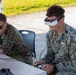 Two U.S. Marines in MARPAT digital camouflage uniforms are seated at a table under a canopy, operating what appears to be a drone or unmanned aerial system. The Marine on the right is wearing FPV (First Person View) goggles branded 'ARK' and holding a remote controller, while the Marine on the left monitors equipment. This appears to be a drone operation or training exercise conducted outdoors.