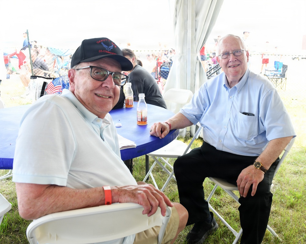 Two elderly men are seated at a blue folding table under a white tent at what appears to be an outdoor public event, possibly an air show. Both men are smiling at the camera. The background shows a crowd of spectators and American flags visible beyond the tent.