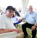 Two elderly men are seated at a blue folding table under a white tent at what appears to be an outdoor public event, possibly an air show. Both men are smiling at the camera. The background shows a crowd of spectators and American flags visible beyond the tent.