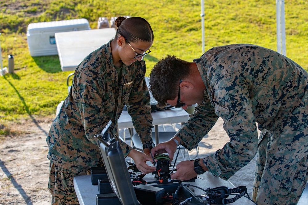 Two U.S. Marines in MARPAT camouflage uniforms are working together to assemble or inspect a small unmanned aerial vehicle (drone) on a table in an outdoor setting. Both personnel are leaning over the equipment, appearing to connect wiring or components on the drone. The setting appears to be a field training or exercise environment with a tent, folding tables, and a cooler visible in the background.