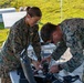 Two U.S. Marines in MARPAT camouflage uniforms are working together to assemble or inspect a small unmanned aerial vehicle (drone) on a table in an outdoor setting. Both personnel are leaning over the equipment, appearing to connect wiring or components on the drone. The setting appears to be a field training or exercise environment with a tent, folding tables, and a cooler visible in the background.