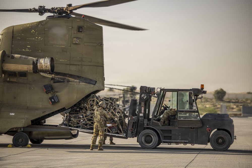 Military personnel are loading or unloading cargo from the rear ramp of a CH-47 Chinook helicopter using a heavy-duty military forklift on a tarmac. The operation appears to take place in an arid, desert-like environment, likely a forward operating base or military airfield. At least two soldiers in camouflage uniforms are actively involved in the cargo handling operation.