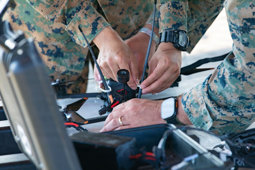 Two individuals wearing US Marine Corps MARPAT digital camouflage uniforms are assembling or preparing a small drone/UAV on what appears to be a portable case or work surface. The drone has visible antennas, a camera mount, and orange-accented frame components. This appears to be a field preparation or maintenance activity involving a small unmanned aerial system.