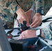Two individuals wearing US Marine Corps MARPAT digital camouflage uniforms are assembling or preparing a small drone/UAV on what appears to be a portable case or work surface. The drone has visible antennas, a camera mount, and orange-accented frame components. This appears to be a field preparation or maintenance activity involving a small unmanned aerial system.