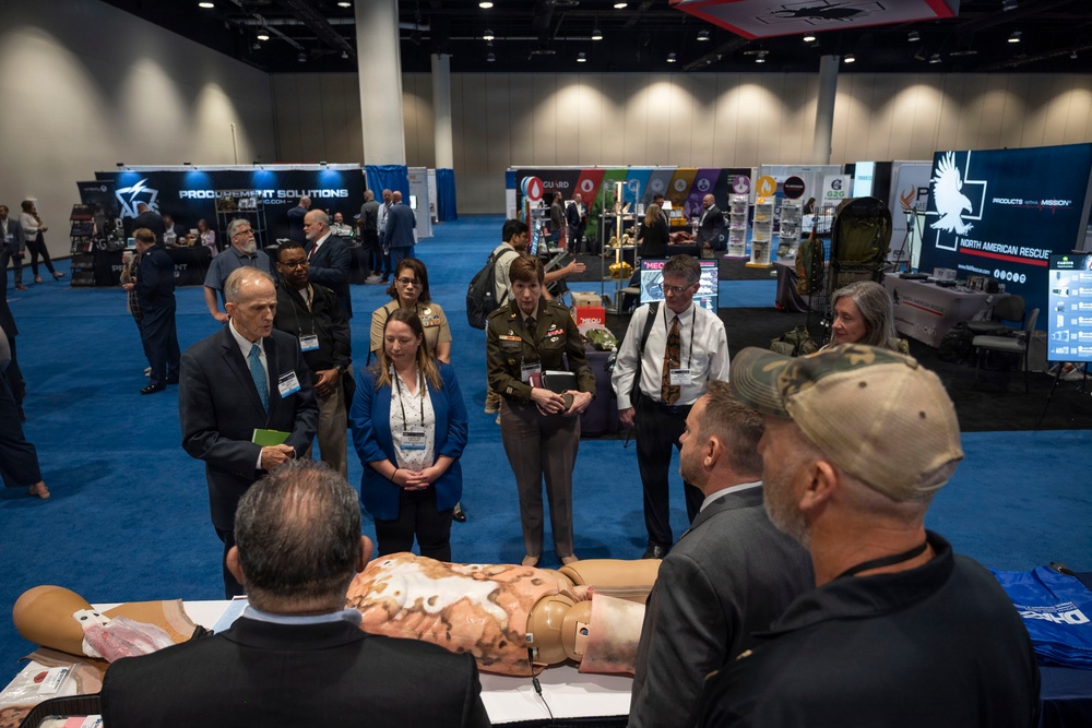 A military and defense trade show or conference is taking place in a large convention center. Attendees, including military personnel in uniform and civilians with badge lanyards, are gathered around a medical training manikin displaying simulated wounds. Vendor booths in the background include 'Procurement Solutions' and 'North American Rescue,' suggesting a focus on military medical or tactical equipment.