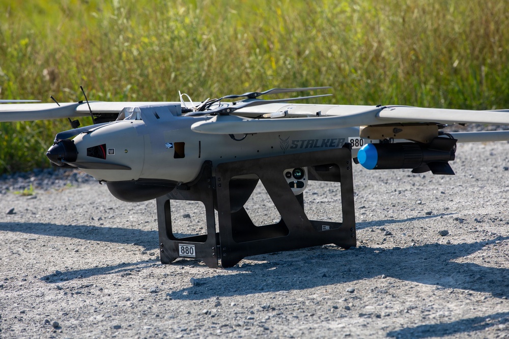 The image shows a fixed-wing UAV (unmanned aerial vehicle) designated 'STALKER 880' resting on a ground launch/support stand on a gravel surface. The drone features a twin-boom pusher configuration with vertical takeoff rotors mounted above the fuselage, suggesting a VTOL (Vertical Take-Off and Landing) hybrid design. The aircraft appears to be in a pre-flight or post-flight ground state in an outdoor field environment.