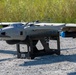 The image shows a fixed-wing UAV (unmanned aerial vehicle) designated 'STALKER 880' resting on a ground launch/support stand on a gravel surface. The drone features a twin-boom pusher configuration with vertical takeoff rotors mounted above the fuselage, suggesting a VTOL (Vertical Take-Off and Landing) hybrid design. The aircraft appears to be in a pre-flight or post-flight ground state in an outdoor field environment.