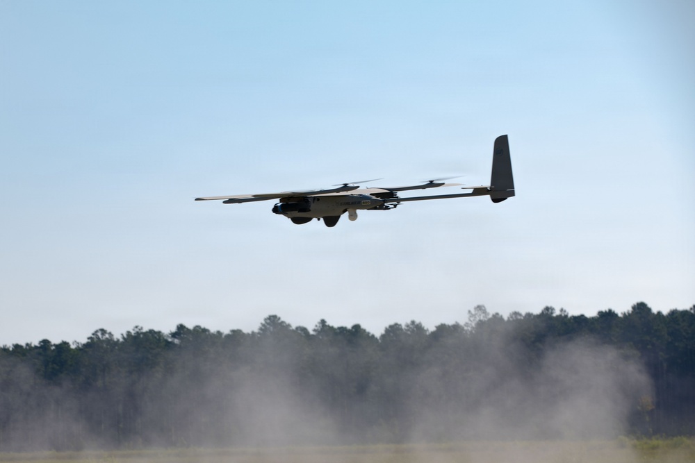 A fixed-wing UAV (unmanned aerial vehicle) with multiple rotors is captured in low-altitude flight over an open field. The aircraft appears to be a hybrid VTOL (Vertical Take-Off and Landing) drone, combining fixed-wing design with multiple electric rotors mounted along the wings. Dust or smoke is visible near the ground below, suggesting a recent takeoff or nearby disturbance.