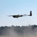 A fixed-wing UAV (unmanned aerial vehicle) with multiple rotors is captured in low-altitude flight over an open field. The aircraft appears to be a hybrid VTOL (Vertical Take-Off and Landing) drone, combining fixed-wing design with multiple electric rotors mounted along the wings. Dust or smoke is visible near the ground below, suggesting a recent takeoff or nearby disturbance.