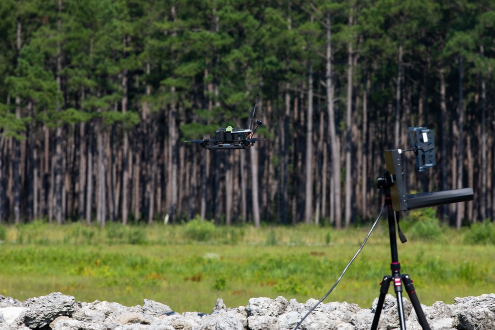 A small quadcopter drone is captured mid-flight in an outdoor setting near a treeline of tall pine trees. On the right side of the frame, a tripod-mounted device — likely a drone detection, tracking, or counter-drone system — is pointed toward the airborne drone. The scene appears to be a controlled test or demonstration environment, possibly military or research-related.