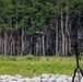 A small quadcopter drone is captured mid-flight in an outdoor setting near a treeline of tall pine trees. On the right side of the frame, a tripod-mounted device — likely a drone detection, tracking, or counter-drone system — is pointed toward the airborne drone. The scene appears to be a controlled test or demonstration environment, possibly military or research-related.