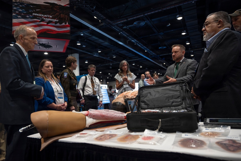 A group of professionals, including military personnel and civilians, are gathered around a display table at what appears to be a defense or medical trade show. A presenter in a gray suit is demonstrating medical simulation or wound care training products, including a prosthetic limb model and packaged wound simulation materials. A banner reading 'Products With a Mission' with an American flag is visible in the background.