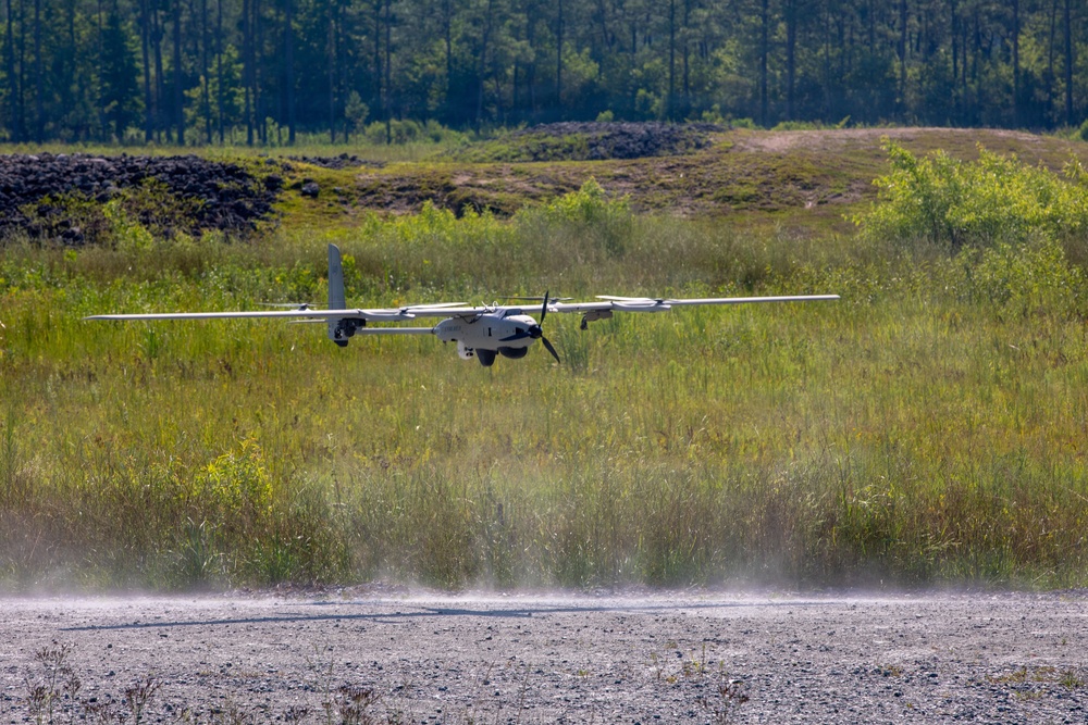 A fixed-wing unmanned aerial vehicle (UAV) is captured at low altitude during takeoff or landing on a gravel/dirt strip, with dust being kicked up beneath it. The aircraft features a pusher propeller configuration, twin-boom tail design, and multiple rotors suggesting a hybrid VTOL (Vertical Take-Off and Landing) capability. The setting is a rural or military training area surrounded by tall grass, scrub vegetation, and a tree line in the background.