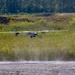 A fixed-wing unmanned aerial vehicle (UAV) is captured at low altitude during takeoff or landing on a gravel/dirt strip, with dust being kicked up beneath it. The aircraft features a pusher propeller configuration, twin-boom tail design, and multiple rotors suggesting a hybrid VTOL (Vertical Take-Off and Landing) capability. The setting is a rural or military training area surrounded by tall grass, scrub vegetation, and a tree line in the background.