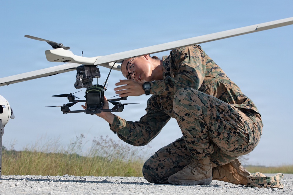 A military service member in MARPAT (Marine pattern) digital camouflage uniform is crouching on a gravel surface while closely inspecting or adjusting a small quadcopter drone attached beneath a larger fixed-wing UAV. The individual is wearing glasses and a watch, and appears to be conducting a pre-flight check or maintenance on the unmanned aerial systems. The setting appears to be an outdoor airfield or training area under clear blue skies.