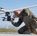 A military service member in MARPAT (Marine pattern) digital camouflage uniform is crouching on a gravel surface while closely inspecting or adjusting a small quadcopter drone attached beneath a larger fixed-wing UAV. The individual is wearing glasses and a watch, and appears to be conducting a pre-flight check or maintenance on the unmanned aerial systems. The setting appears to be an outdoor airfield or training area under clear blue skies.