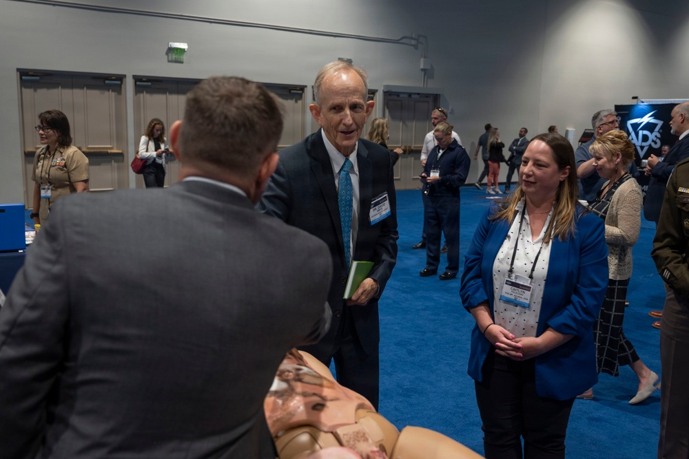 A professional conference or trade show event taking place in a large convention hall with blue carpeting. Attendees wearing name badges are engaged in networking conversations, with exhibition booths visible in the background including one marked 'ADS'. The foreground shows a group of three people in business attire engaged in conversation.