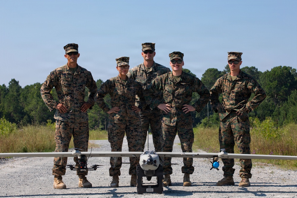 Five military personnel in MARPAT camouflage uniforms pose together on a gravel path in front of unmanned aerial vehicles (UAVs). The group includes both male and female service members standing with hands on hips in a confident posture. Two distinct UAV systems are visible in the foreground, including a fixed-wing drone marked '880' and a smaller quadcopter.