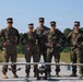 Five military personnel in MARPAT camouflage uniforms pose together on a gravel path in front of unmanned aerial vehicles (UAVs). The group includes both male and female service members standing with hands on hips in a confident posture. Two distinct UAV systems are visible in the foreground, including a fixed-wing drone marked '880' and a smaller quadcopter.