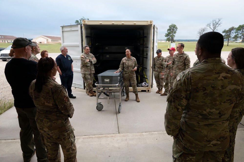 A group of military personnel in camouflage uniforms, along with a few civilians, are gathered in a semicircle outdoors near a large open shipping container. A female service member appears to be briefing or instructing the group while standing next to a wheeled metal cart or case. The setting appears to be a military installation based on the buildings and infrastructure visible in the background.