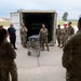 A group of military personnel in camouflage uniforms, along with a few civilians, are gathered in a semicircle outdoors near a large open shipping container. A female service member appears to be briefing or instructing the group while standing next to a wheeled metal cart or case. The setting appears to be a military installation based on the buildings and infrastructure visible in the background.