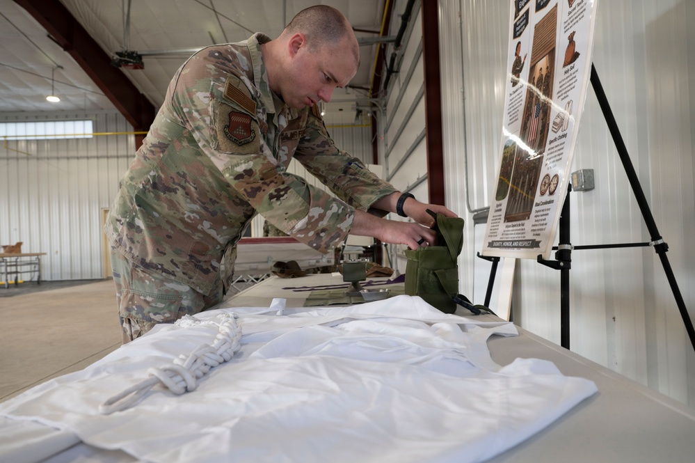 A U.S. Air Force service member in OCP (Operational Camouflage Pattern) uniform is working at a table in what appears to be a hangar or large indoor facility. He is handling what appears to be military gear or equipment laid out on a white surface. An instructional poster on an easel is visible in the background, with text referencing 'Dignity, Honor and Respect.'