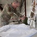A U.S. Air Force service member in OCP (Operational Camouflage Pattern) uniform is working at a table in what appears to be a hangar or large indoor facility. He is handling what appears to be military gear or equipment laid out on a white surface. An instructional poster on an easel is visible in the background, with text referencing 'Dignity, Honor and Respect.'