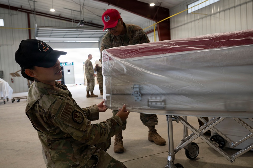 Two military personnel in OCP (Operational Camouflage Pattern) uniforms are working with a flag-draped casket wrapped in protective plastic sheeting inside what appears to be a military hangar. The casket, which displays red, white, and blue colors consistent with an American flag, is positioned on a wheeled transfer case. This appears to be a dignified transfer or mortuary affairs operation.
